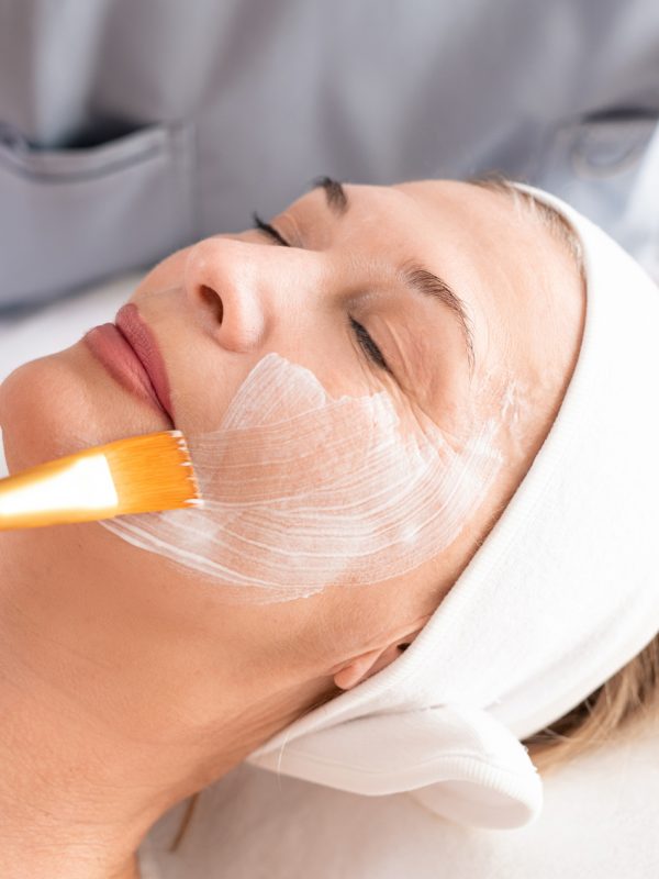 Close-up of beauty professional applying facial mask to relaxed mature woman with closed eyes at spa procedure